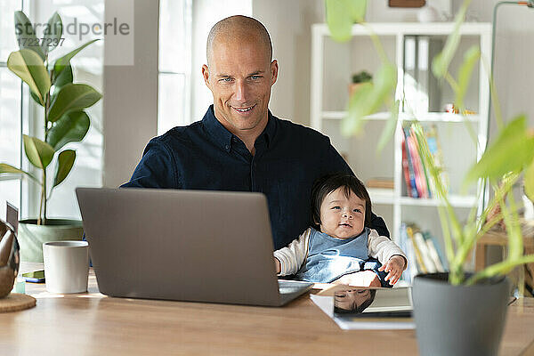 Vater arbeitet am Laptop  während er mit seinem Baby im Heimbüro sitzt