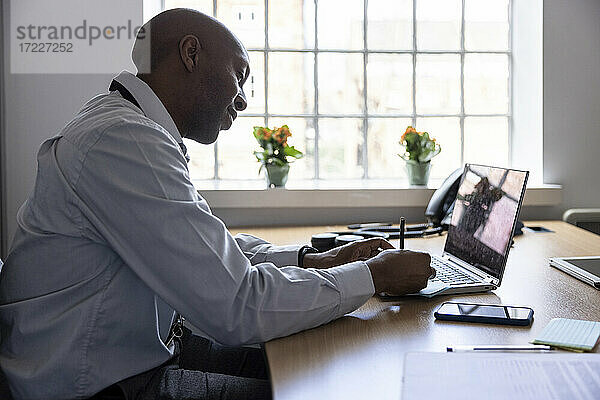Männlicher Berufstätiger  der auf einem Laptop eine Haftnotiz schreibt  während er am Schreibtisch im Büro sitzt