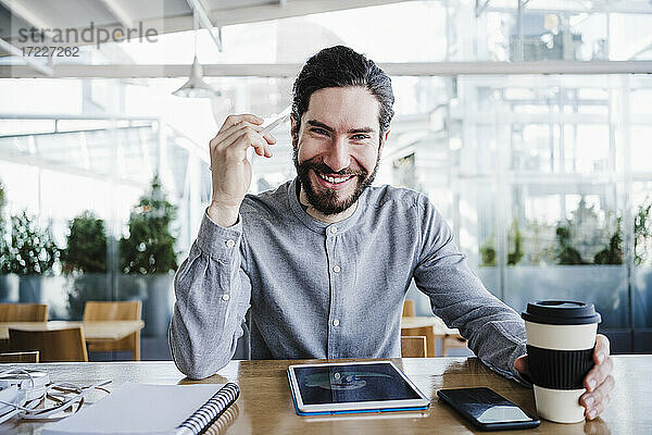 Lächelnder männlicher Freiberufler mit wiederverwendbarem Kaffeebecher durch drahtlose Technologien im Büro