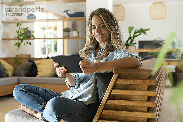 Lächelnde Frau  die ein Video über ein digitales Tablet ansieht  während sie auf der Couch im Wohnzimmer sitzt