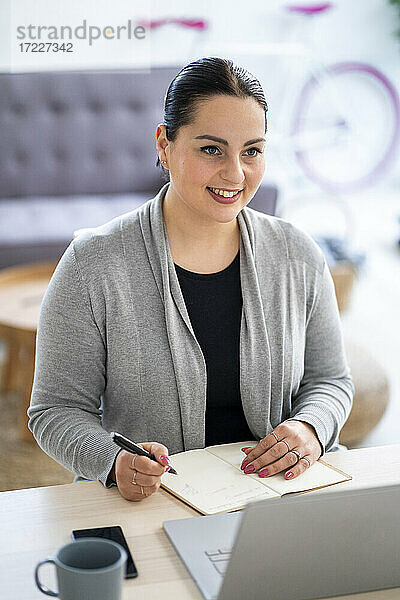 Lächelnde Geschäftsfrau  die einen Laptop benutzt und in ihr Tagebuch schreibt  während sie zu Hause arbeitet