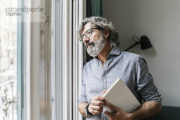 Nachdenklicher älterer Mann hält ein Buch in der Hand  während er durch das Fenster seines Hauses schaut