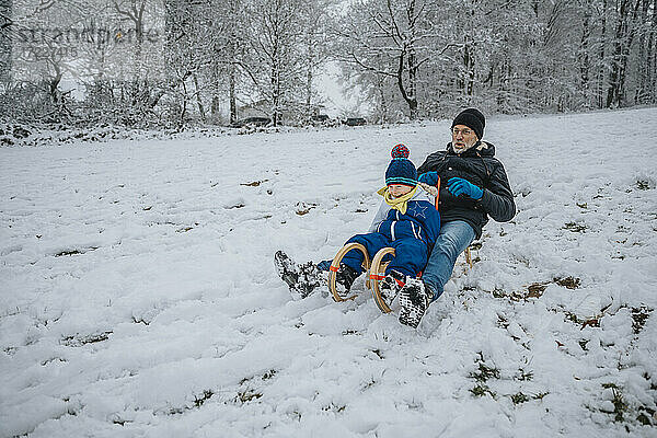 Verspielter Vater und Sohn beim Schlittenfahren auf einem verschneiten Hügel im Winter