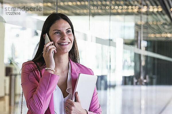 Lächelnde Geschäftsfrau mit Laptop  die vor einer Glaswand mit einem Mobiltelefon spricht