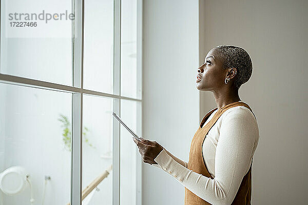 Nachdenkliche Frau  die ein digitales Tablet hält  während sie zu Hause vor einem Fenster steht