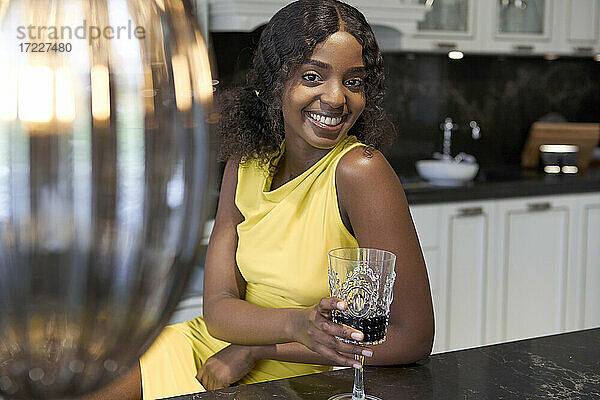 Porträt einer glücklichen jungen Frau mit einem Glas Rotwein in der Küche sitzend