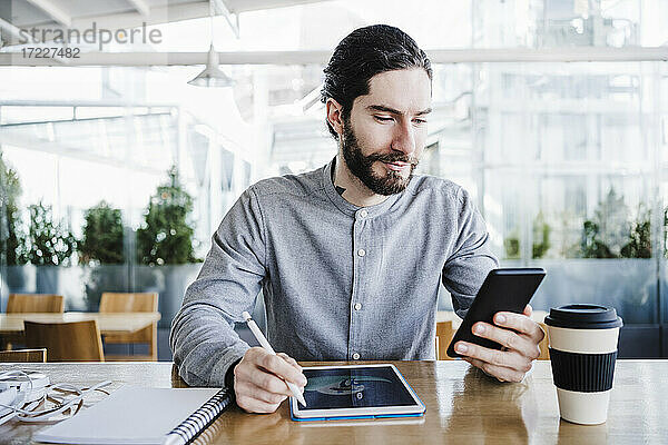 Geschäftsmann mit Grafiktablett und wiederverwendbarem Kaffeebecher bei der Nutzung eines Smartphones im Büro