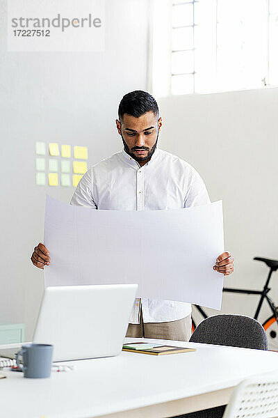 Männlicher Unternehmer  der im Büro stehend einen Geschäftsplan am Laptop hält