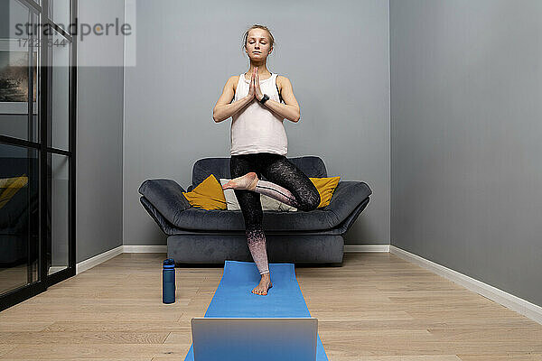Junge Frau macht Yoga  während sie mit ihrem Laptop auf einer Übungsmatte zu Hause steht