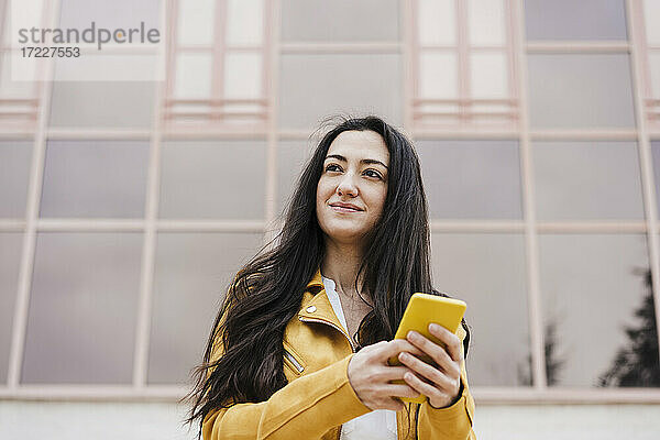Lächelnde Frau  die wegschaut  während sie ein Mobiltelefon vor einem Gebäude benutzt