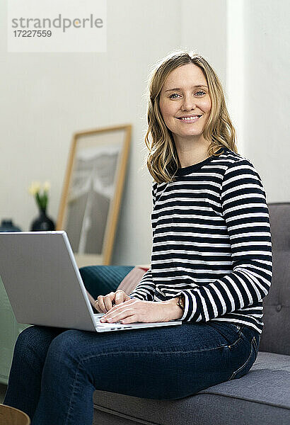Lächelnde blonde Frau sitzt mit Laptop auf dem Sofa zu Hause