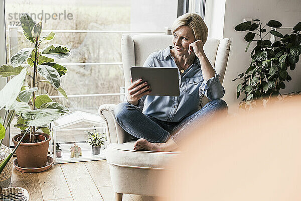 Kontemplative Frau mit digitalem Tablet  die im Wohnzimmer wegschaut