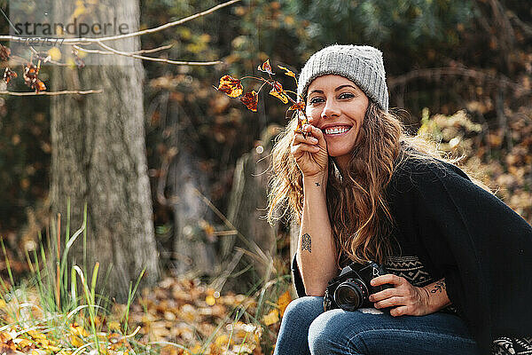 Lächelnde schöne Frau  die Blätter hält  während sie mit der Kamera im Wald im Herbst sitzt