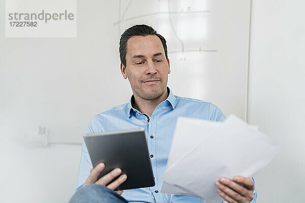 Männlicher Unternehmer  der ein digitales Tablet und Dokumente hält  während er im Büro sitzt