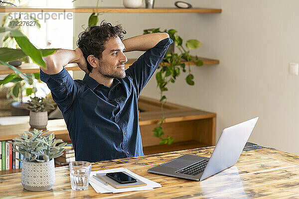 Entspannter männlicher Freiberufler mit Händen hinter dem Kopf  der wegschaut  während er vor einem Laptop am Tisch sitzt