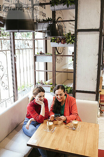 Lächelnde Frau  die ein Smartphone benutzt  während sie neben einer Freundin mit der Hand am Kinn in einem Café sitzt