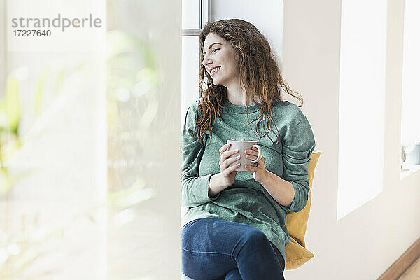 Lächelnde junge Frau mit Kaffeetasse  die nachdenklich am Fenster im Wohnzimmer sitzt