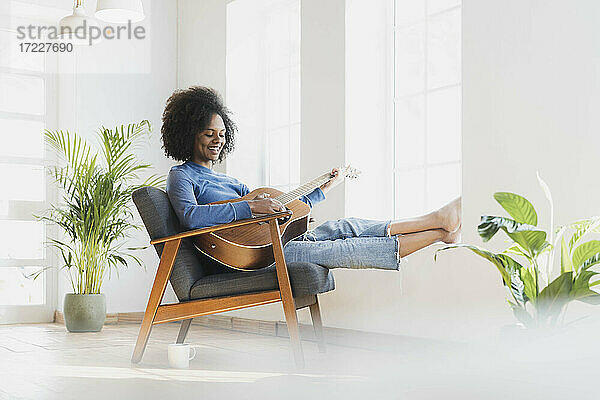 Lächelnde junge Frau spielt Gitarre  während sie zu Hause auf einem Sessel sitzt
