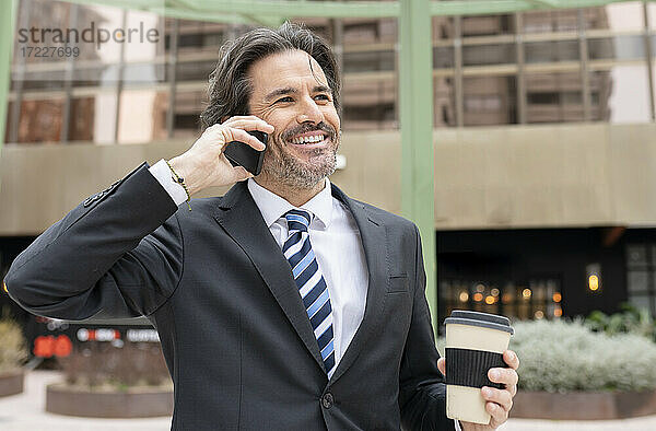 Lächelnder Geschäftsmann mit Kaffeetasse  der im Büropark mit seinem Handy telefoniert