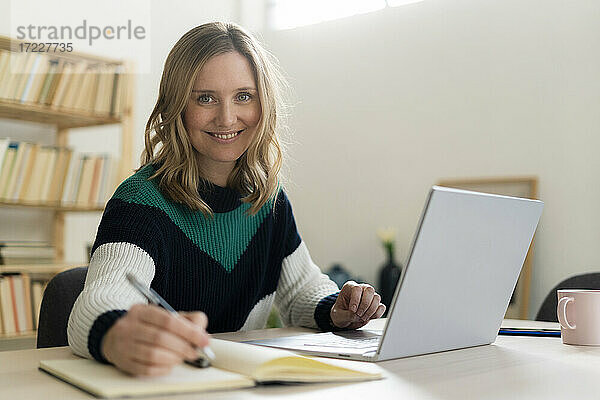 Blonde Frau lächelt  während sie zu Hause an ihrem Laptop sitzt