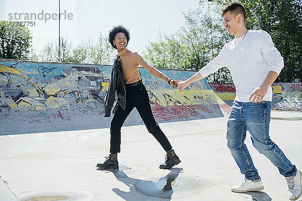 Verspieltes junges Paar  das sich in einem Skateboard-Park an einem sonnigen Tag an den Händen hält
