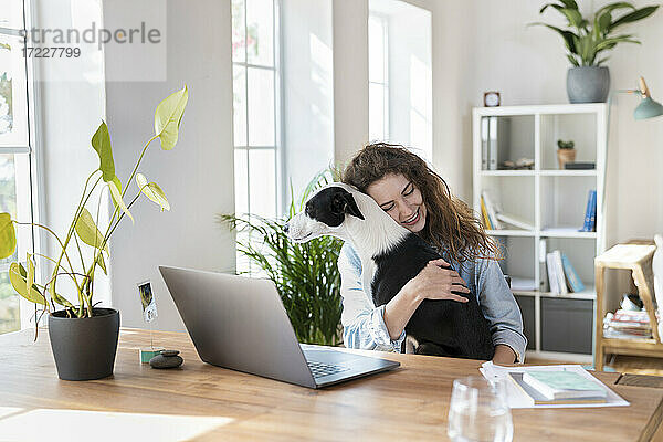 Lächelnde weibliche Fachkraft mit geschlossenen Augen  die einen Jack Russell Terrier im Büro zu Hause umarmt