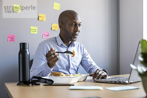Männlicher Berufstätiger isst Salat und schaut auf einen Laptop im Büro