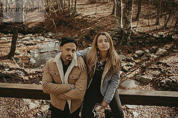 Hipster-Mann steht neben seiner Freundin  die auf einem Holzgeländer im Wald sitzt  im Herbst