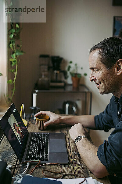 Lächelnder Geschäftsmann in Diskussion über Videoanruf mit männlichem Kollegen durch Laptop während Pandemie