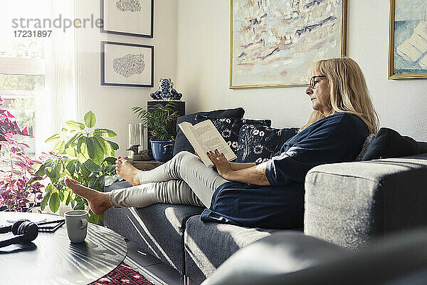 Blonde Frau liest Buch  während sie auf dem Sofa im Wohnzimmer sitzt