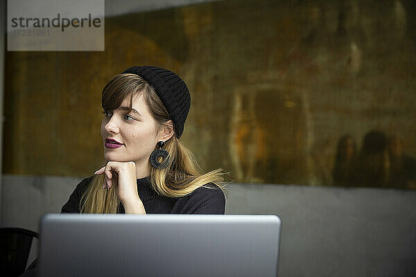 Lächelnde Geschäftsfrau mit Hand am Kinn durch Laptop im Büro