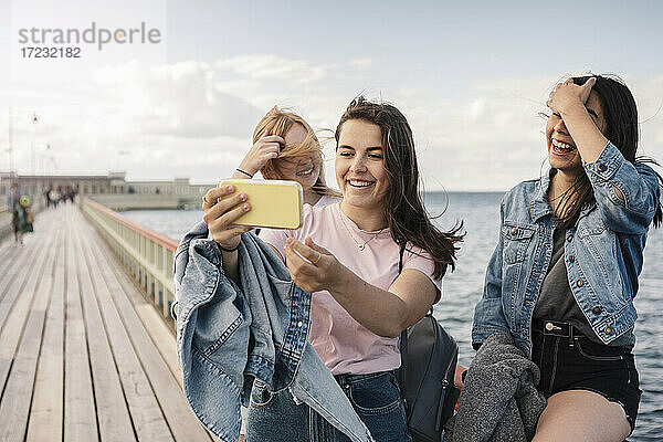 Glückliche weibliche Freunde nehmen Selfie durch Smartphone auf Pier gegen Meer