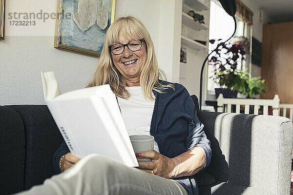 Lächelnde Frau  die ein Buch liest  während sie auf dem Sofa zu Hause sitzt