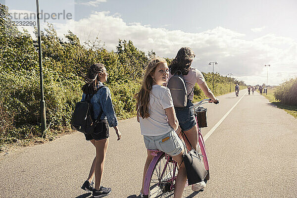 Weibliche Freunde Radfahren während Teenager-Mädchen zu Fuß auf der Straße während sonnigen Tag gegen Himmel