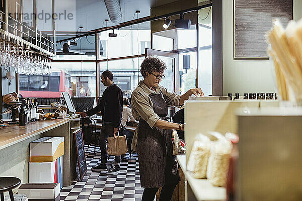 Weibliche Ladenbesitzerin bei der Arbeit im Feinkostladen mit Kunden im Hintergrund