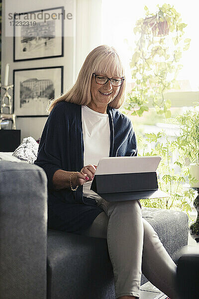 Lächelnde Frau mit digitaler Tablette beim Sitzen auf dem Sofa zu Hause