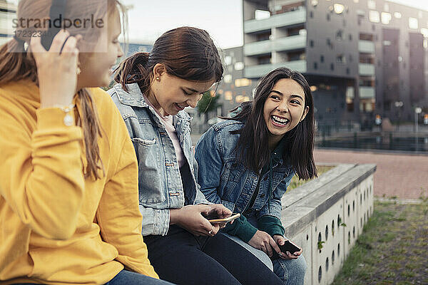 Glückliche weibliche Freunde mit Smartphone während des Gesprächs mit einander an Rückhaltewand