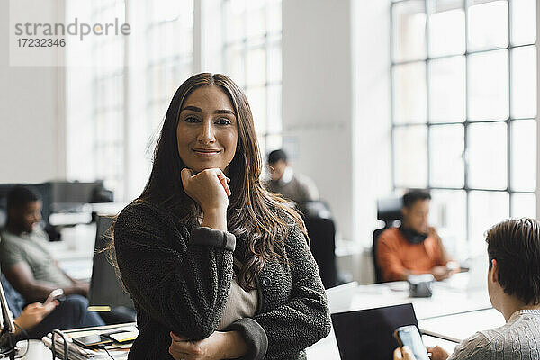 Lächelnde Unternehmerin mit Hand am Kinn im kreativen Büro