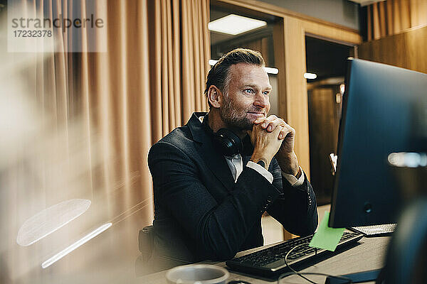 Lächelnde männliche Unternehmer mit Hand auf Kinn arbeiten über Laptop im Büro