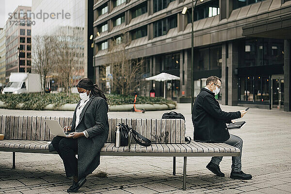 Geschäftsmann mit Smartphone  während eine Kollegin mit Laptop auf einer Bank sitzt  während COVID-19