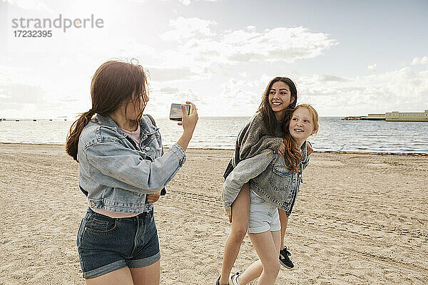 Teenager-Mädchen geben Huckepack Fahrt  während weibliche Freunde am Strand gegen Meer und Himmel zu fotografieren