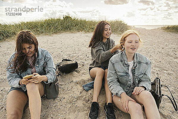 Teenager-Mädchen mit Smartphone während weibliche Freunde sitzen auf Sand am Strand