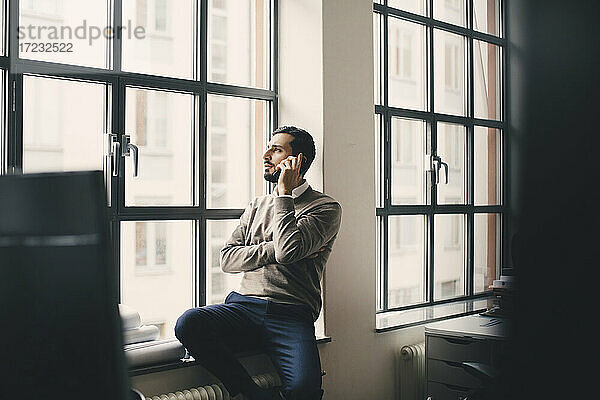 Männlicher Unternehmer  der am Fenster in einem kreativen Büro mit einem Smartphone spricht