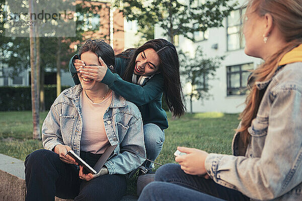 Lächelnde Freundinnen decken die Augen eines Teenagers ab  während sie auf einer Stützmauer sitzen