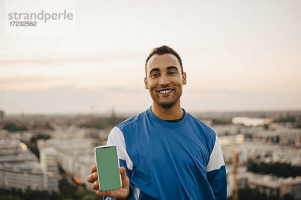 Lächelnde männliche Sportler hält Smartphone gegen den Himmel während des Sonnenuntergangs
