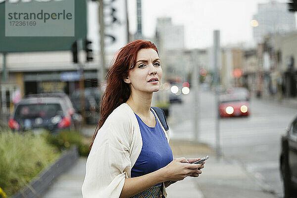 Junge Frau mit langen roten Haaren  mit Smartphone auf der Straße