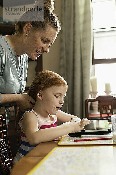 Mädchen mit digitalem Tablet am Esstisch  während die Mutter ihr Haar stylt