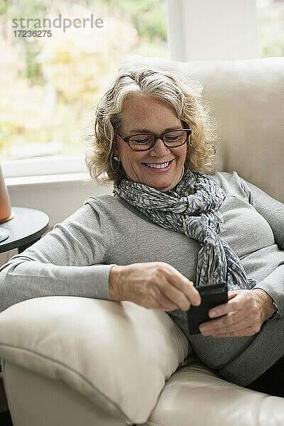 Entspannte ältere Frau sitzt auf dem Sofa und benutzt ein Mobiltelefon