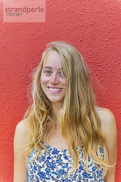 Portrait einer Jungen Frau mit langen blonden Haaren vor einer roten Wand  lacht  Insel Burano  Venedig  Venetien  Italien  Europa