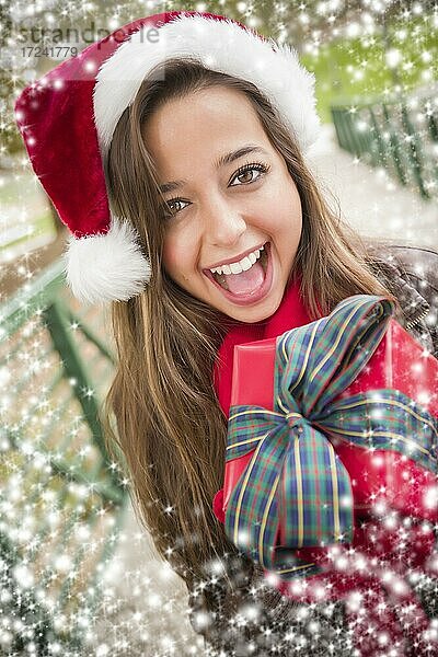 Hübsche festlich gekleidete lächelnde Frau trägt einen Weihnachtsmann Hut eingewickelt Geschenk draußen mit Schneeflocken